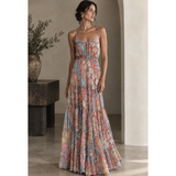 A woman models a sleeveless, tiered maxi dress with a colorful floral and paisley print in shades of pink, blue, orange, and beige, set against a warm-toned architectural background.