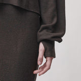 Close-up of a person wearing a dark brown knit garment with a gathered sleeve and long cuff, shown against a plain light gray background.