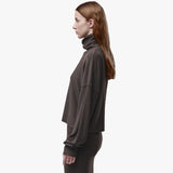 A woman in side profile wearing a dark brown cropped turtleneck sweatshirt and matching dark pants, photographed against a clean white studio background.