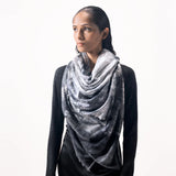 A woman wearing a limited edition hand-painted cashmere and silk scarf in gray, black, and white tones with frayed edges and a silver lamination.
