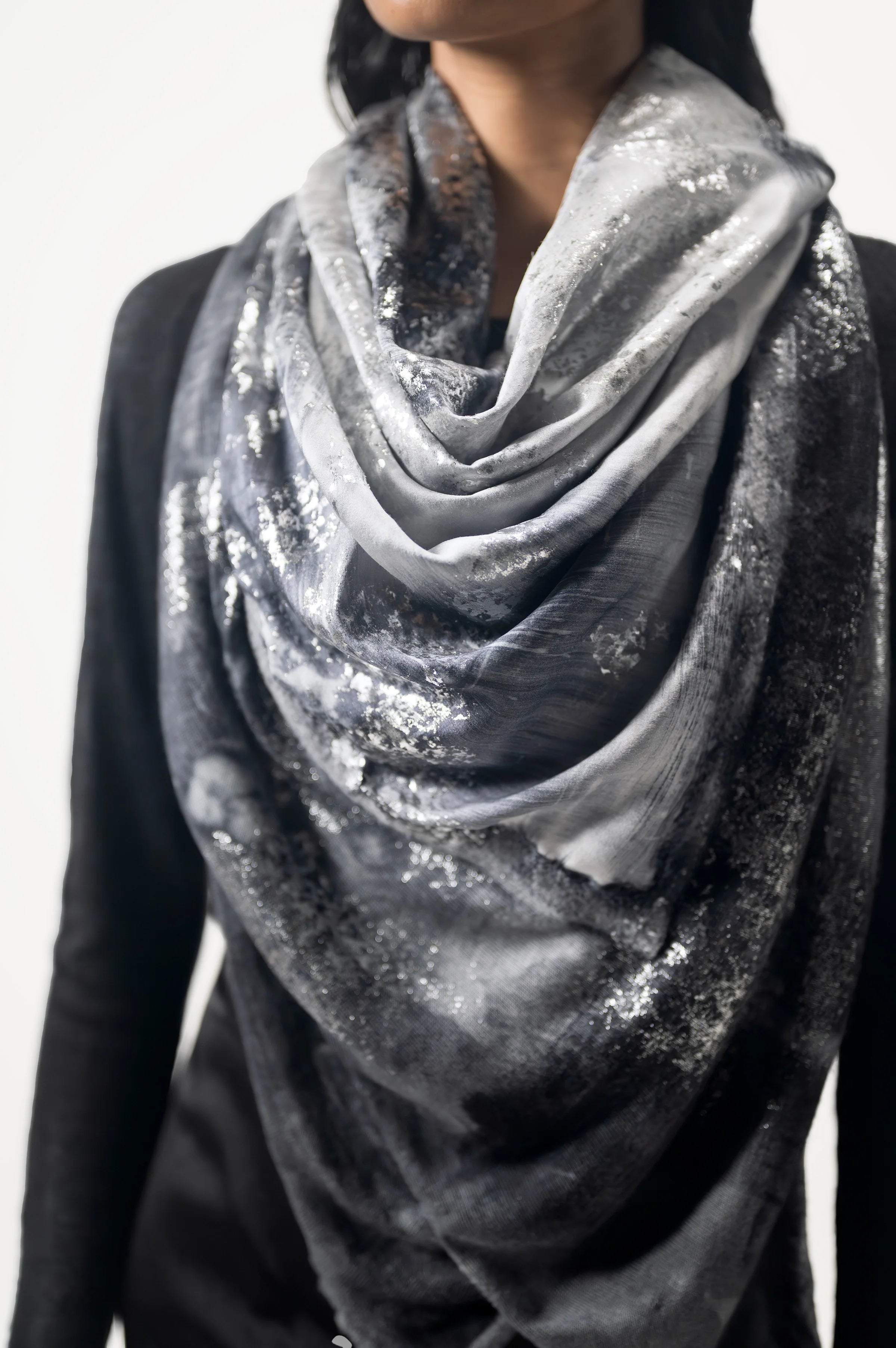 A woman wearing a limited edition hand-painted cashmere and silk scarf in gray, black, and white tones with frayed edges and a silver lamination.