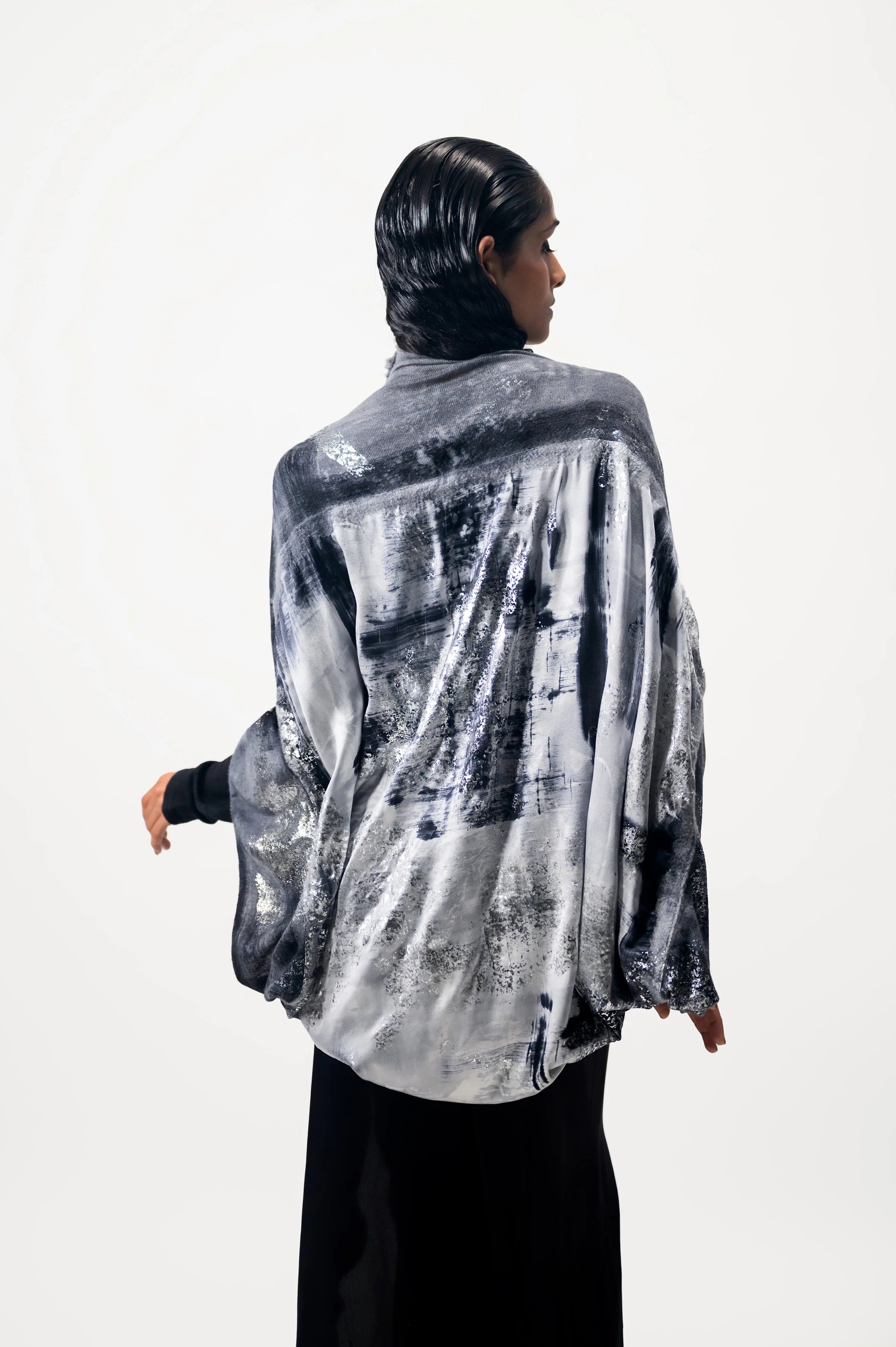 A woman wearing a limited edition hand-painted cashmere and silk scarf in gray, black, and white tones with frayed edges and a silver lamination.